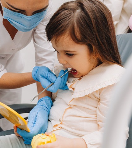 Little girl in dental chair looking into handheld mirror while dentist examines the backs of her teeth with dental mirror